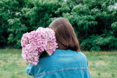 Milenyum kızı kot ceket giyip elinde bir demet taze mor ortanca çiçeğiyle dikiz aynasından bakıyor. Mesaj için yer var. Yaz buketi.