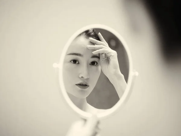 beautiful young asian woman looking at self in mirror, hand on forehead, black and white, retro style.