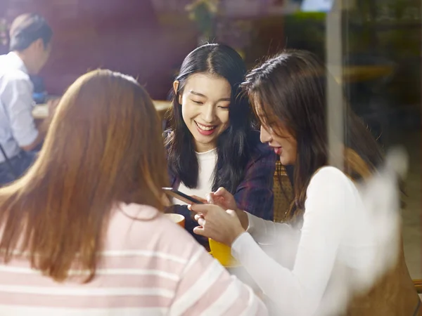 三愉快的美丽的年轻亚洲妇女坐在桌边聊天在咖啡馆或茶馆里玩手机聊天 通过玻璃窗玻璃 — 图库照片