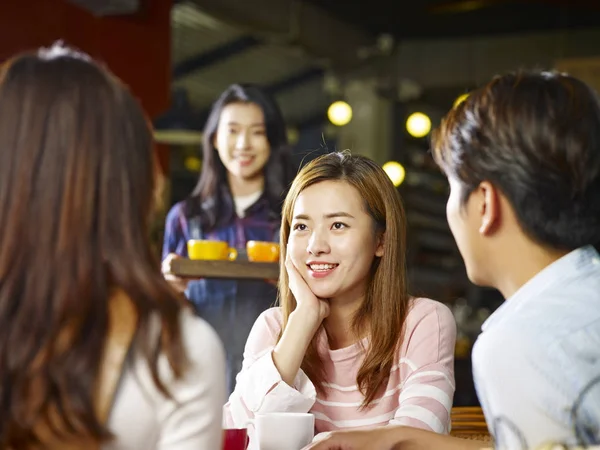 三快乐的亚洲年轻人坐在桌边聊天 当服务生服务咖啡 聚焦前景女孩 — 图库照片