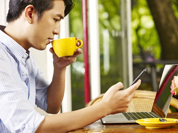 年轻的亚洲男子使用手机和笔记本电脑 同时饮用卡布奇诺咖啡户外 — 图库照片