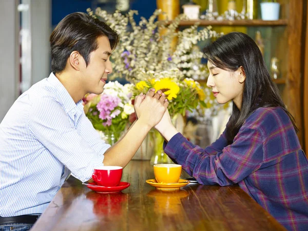 young asian couple sitting face to face holding hands making a wish with eyes closed.