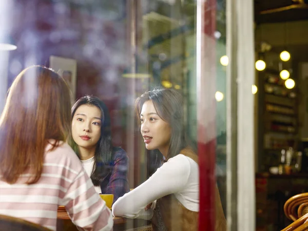 三快乐美丽的年轻亚洲妇女坐在桌边聊天在咖啡馆或茶馆 通过窗口玻璃拍摄 — 图库照片