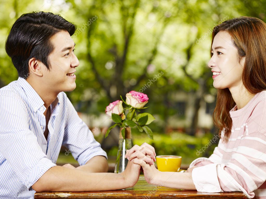 young asian man and woman sitting face to face holding hands looking at each other in coffee shop.