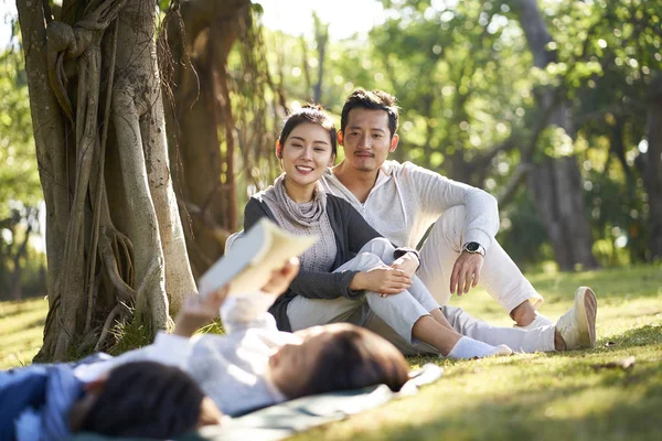 两个亚洲孩子小男孩和女孩有乐趣躺在草地上看书与父母坐在幕后观看 — 图库照片