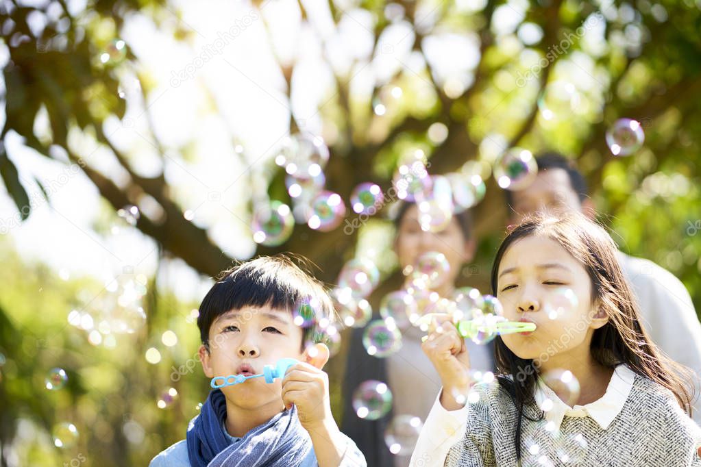 little asian kids boy and girl sister and brother blowing bubbles in a park with parents watching from behind.