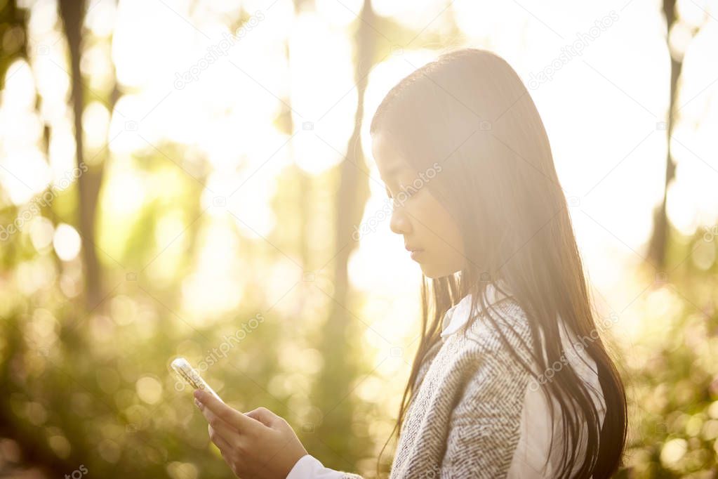 beautiful little asian girl with long hair looking at mobile phone in morning sunlight.