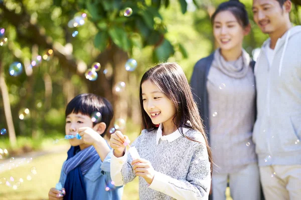 小亚洲孩子男孩和女孩的妹妹和兄弟吹泡泡在公园里 父母从后面看 — 图库照片