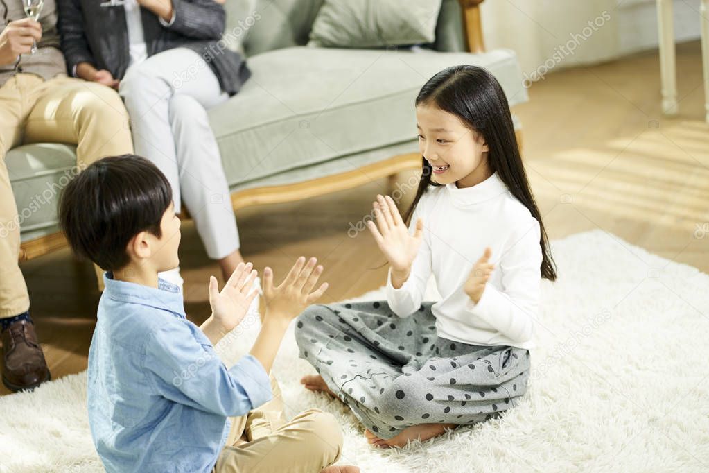 two cute little asian kids brother and sister sitting on carpet playing game with parent sitting on sofa in the background