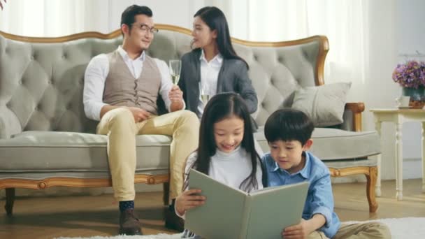 床の上のカーペットの上に座っている2人のアジアの子供たちは 後ろから見ている母と父と一緒に本を読みます — ストック動画