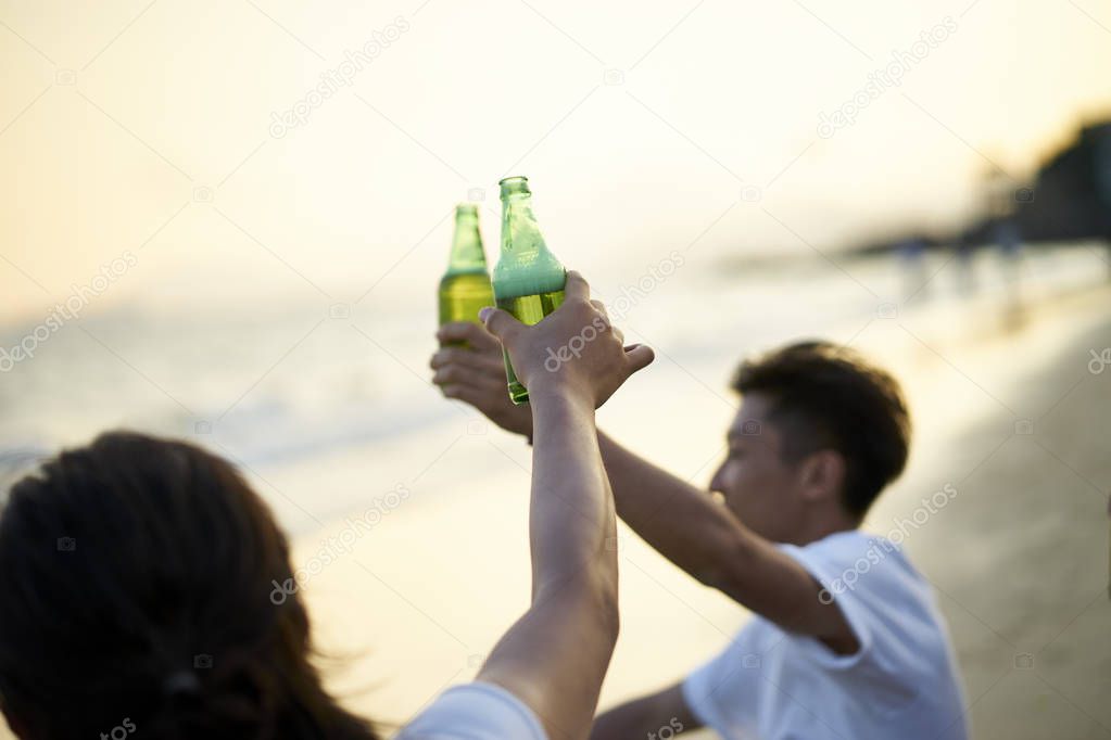 two young asian adult men drinking beer clicking bottles toasting on beach