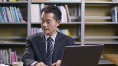 Asyalı şirket yöneticisi iş teklifini okurken dizüstü bilgisayar kullanıyor. 