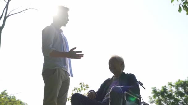 アジア系の大人の息子と車椅子に縛られたお父さんが屋外でおしゃべり — ストック動画