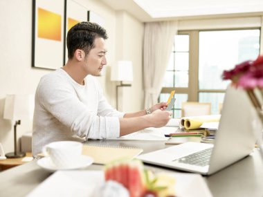 Genç Asyalı tasarım uzmanı mutfakta oturmuş iki renkli kartı karşılaştırıyor (arka planda dijital olarak değiştirilmiş sanat eserleri))