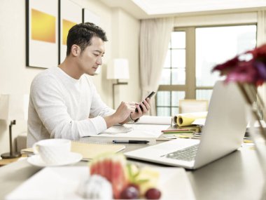 Genç Asyalı tasarım uzmanı mutfakta cep telefonuyla oturuyor (arka planda dijital olarak değiştirilmiş çizimler))