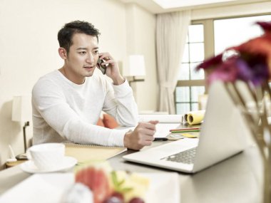 Genç Asyalı tasarım uzmanı evde çalışıyor mutfakta oturuyor ve cep telefonuyla konuşuyor.
