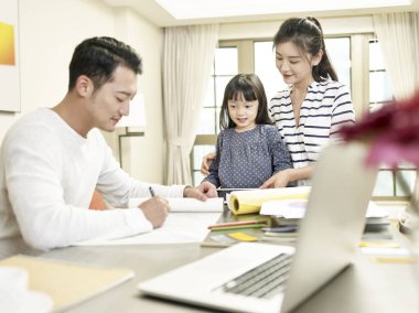 Genç Asyalı anne ve kızı, evde çalışırken babasının bir tasarım çizimini izliyorlar (arka plandaki sanat eserleri dijital olarak değiştirilmiş)