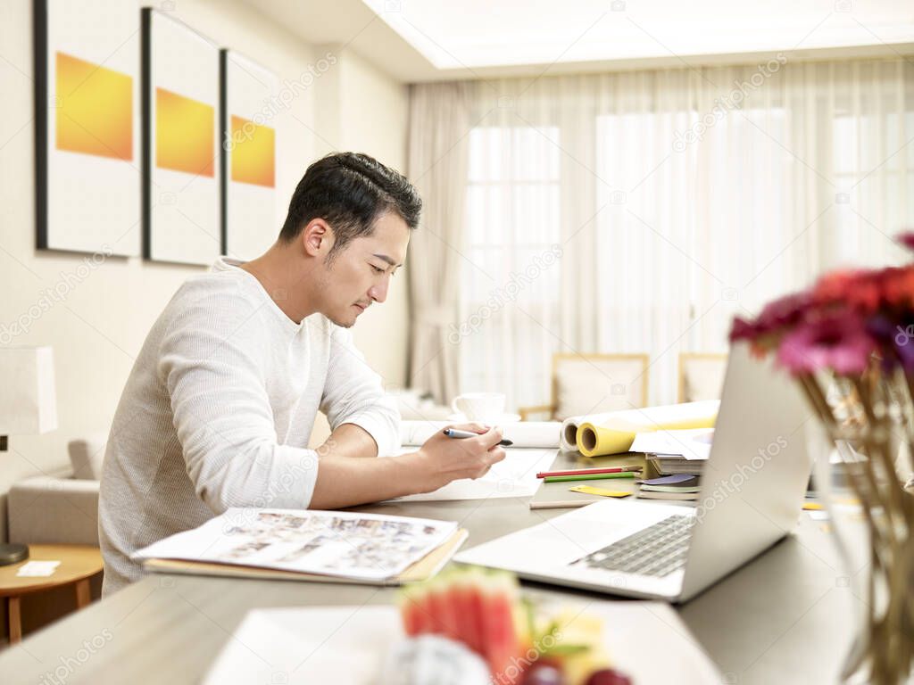 young asian man design professional sitting at kitchen counter working from home (artwork in background digitally altered)