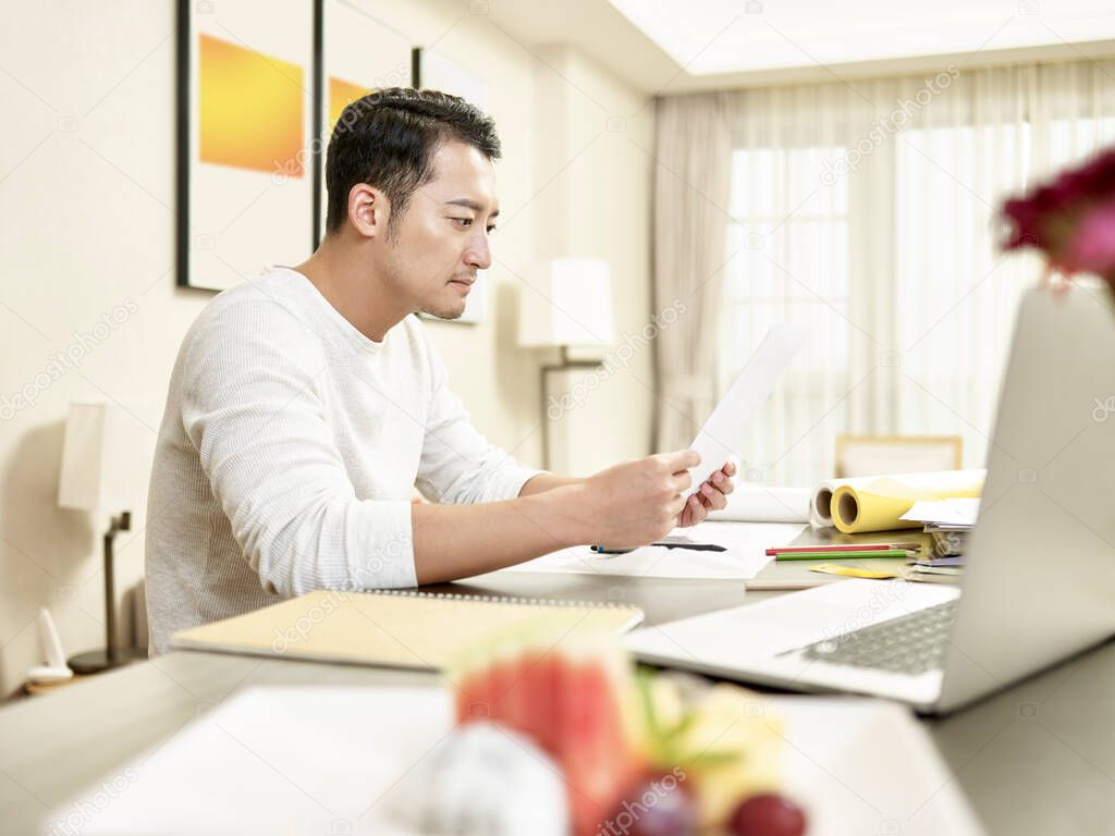 young asian man design professional working from home sitting at kitchen counter reading a document (artwork in background digitally altered)