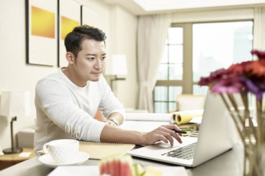 Genç Asyalı işadamı mutfak tezgahında oturmuş dizüstü bilgisayara bakıyor.)