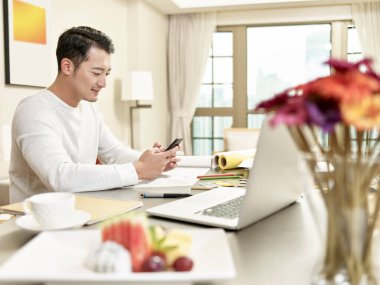 Genç Asyalı tasarım uzmanı mutfakta cep telefonuyla oturuyor (arka planda dijital olarak değiştirilmiş çizimler))