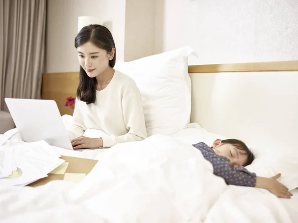 年轻的亚洲母亲在床上使用笔记本电脑 女儿睡在身边 — 图库照片