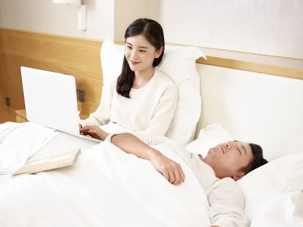 Young Asian Mother Working Using Laptop Computer Bed Husband Sleeping — Stock Photo, Image