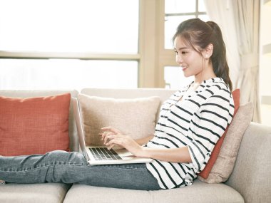 Genç Asyalı kadın evde çalışıyor. Kanepeye oturmuş dizüstü bilgisayar kullanıyor.