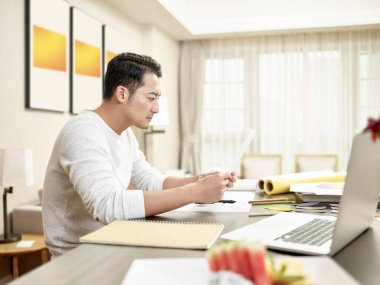 Genç Asyalı tasarım uzmanı mutfak tezgahında oturup bir belge okuyor (dijital olarak değiştirilmiş sanat eserleri))