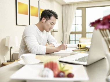 Genç Asyalı tasarım uzmanı mutfak tezgahında oturuyor evden çalışıyor (arka planda dijital olarak değiştirilmiş sanat eseri))