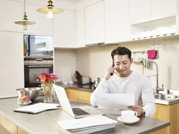 年轻的亚洲商人坐在厨房柜台边 一边在家工作一边看报告 一边用手机讲话 — 图库照片