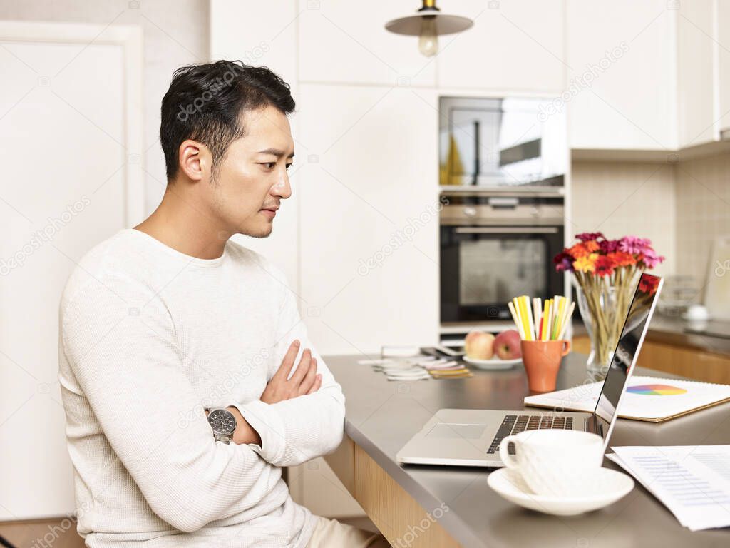 young asian businessman working from home sitting at kitchen counter looking at laptop computer arms crossed