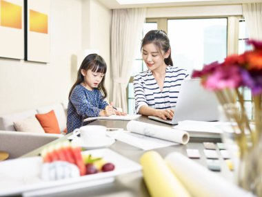 Genç Asyalı tasarımcı anne, kızına bakarken dizüstü bilgisayar kullanıyor (arka planda dijital olarak değiştirilmiş sanat eserleri))