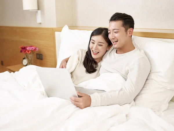 young asian couple sitting in bed enjoying a movie using laptop computer happy and smiling