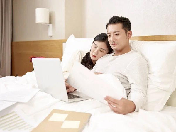 아시아인 사업가가 침대에 앉아서 노트북 컴퓨터를 사용하며 일하고 아내는 옆에서 — 스톡 사진