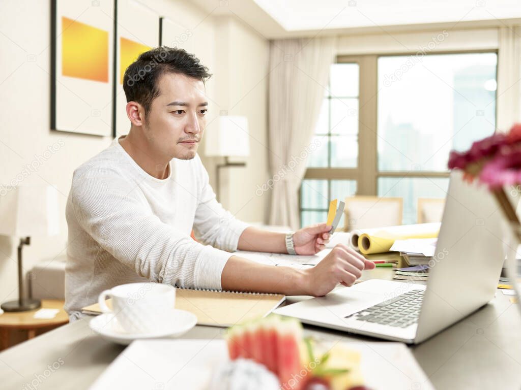 young asian man design professional working from home sitting at kitchen using laptop computer (artwork in background digitally altered)