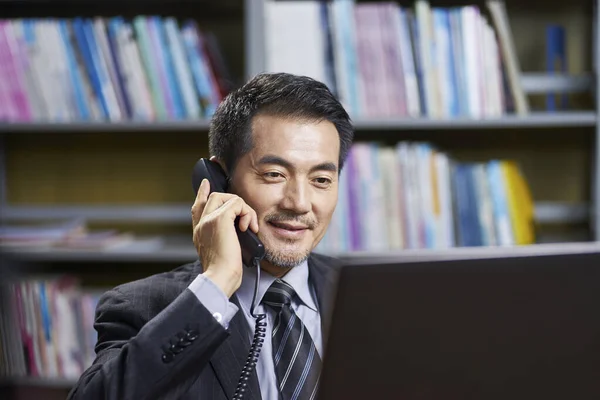 asian corporate executive sitting at desk in office conversing on phone