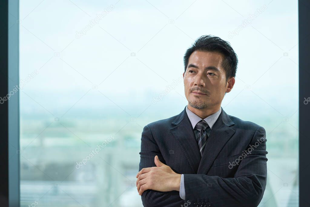 portrait of a mature asian business man standing by window in office arms crossed