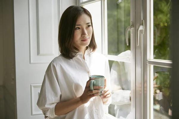 年轻的亚洲女人站在窗边 手里拿着一杯咖啡 — 图库照片