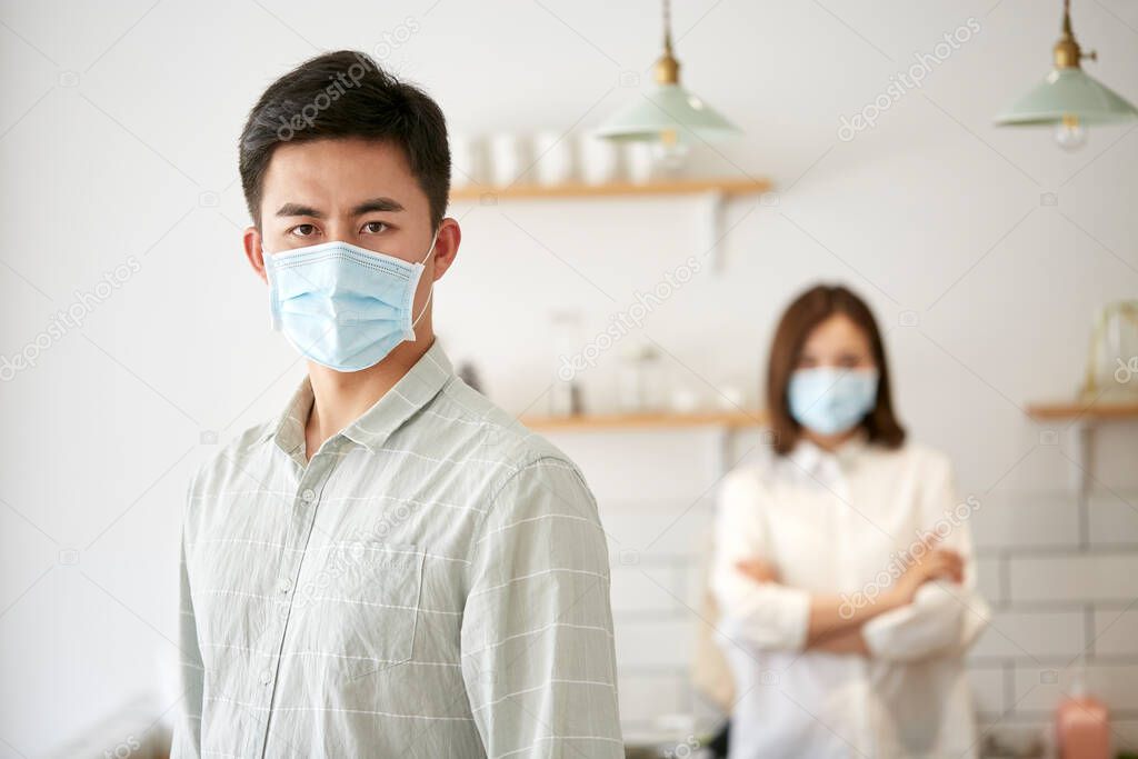 portrait of a young asian man wearing mask looking at camera with husband in the background
