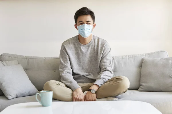 young asian man staying at home wearing mask sitting on couch leg crossed looking at camera