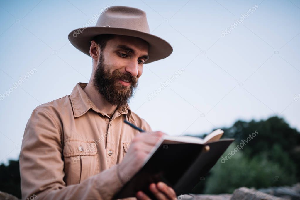 Smiling bearded man wearing hipster hat writing notes in notepad, planning travel. Attractive traveller admire beautiful scenery at sunset. Inspirational travel concept.  