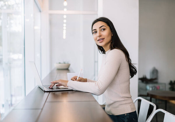 Attractive Indian woman using laptop computer, writing notes, typing on keyboard, working from home. University student learning language, exam preparation. Online education concept