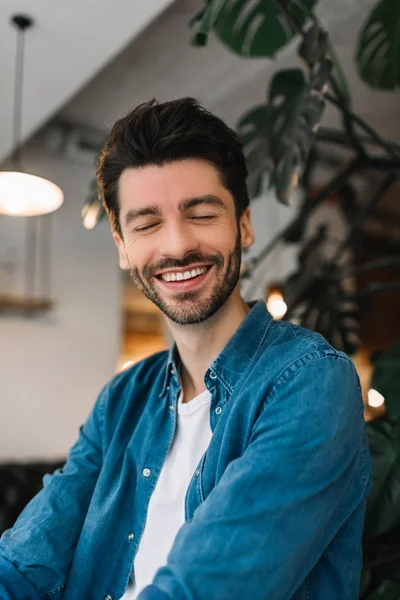 Authentic and natural portrait photography of bearded hipster man with toothy smile and fashion hairstyle sitting in loft modern coffee shop. Satisfied customer wearing casual clothes posing for pictures, good mood, feeling happy.
