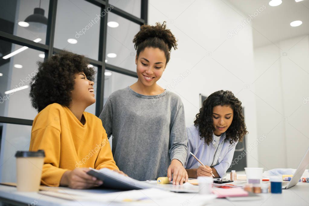 Creative team of smiling African American business women  planning new start up project in creative office. Brainstorming. Collaborate. Talented university students learning fashion design at atelier. Young positive hipsters friends working together.