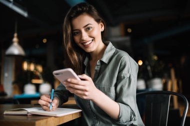 Loft kafede oturan dil öğrenme Web, alışveriş cep telefonuyla çevrimiçi bankacılık, neşeli bir kadın. Eğitim için modern smartphone tutarak ve notlar olumlu beyaz öğrenci. Online eğitim. 