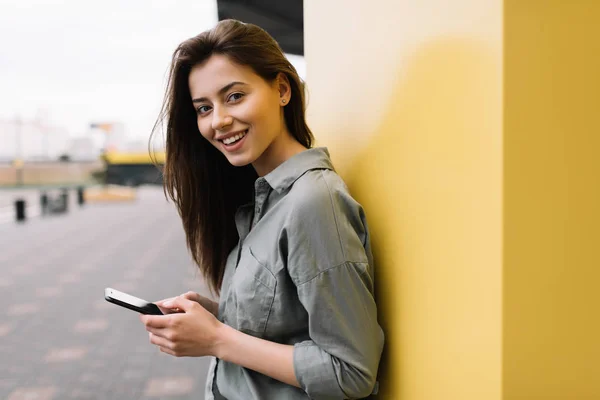 Smartphone duran sarı arka plana yakınındaki kullanan güzel ve olumlu blogger kadın. Müzik, online alışveriş, tipik haber, blog yazısı, resimler için poz indirme hipster giyiyor rahat kıyafetler gülümseyen.