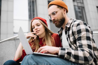 Sakallı adam turuncu şapka ve birlikte çalışan güzel beyaz kadın giyiyor. Şık hipsters arkadaşlar bilgi online araştırma dizüstü bilgisayar kapalı oturan kullanarak. Ekip çalışması. Beyin fırtınası.