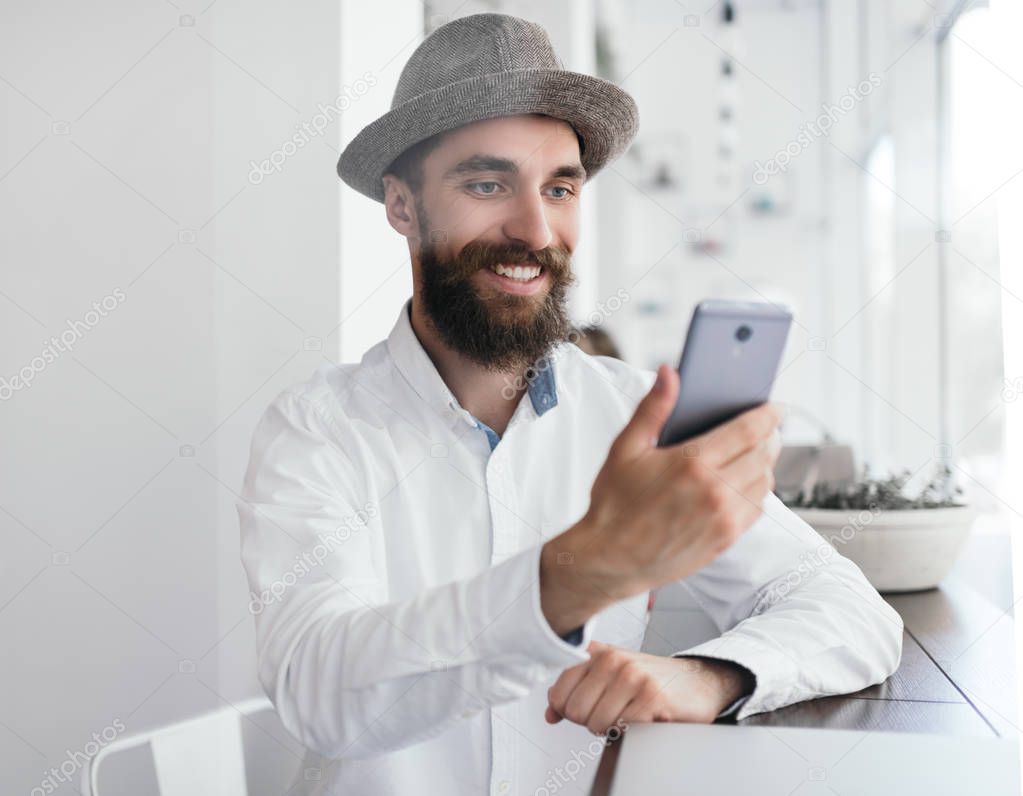 Bearded man with positive and smiling face holding mobile phone, sitting in white loft cafe near window. Attractive stylish blogger streaming video online using smartphone and internet.