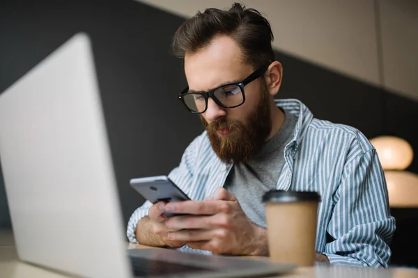 Bearded man wearing hipster glasses using smartphone and laptop computer for his project, downloading application, researching information online. Young caucasian freelancer working in modern office.